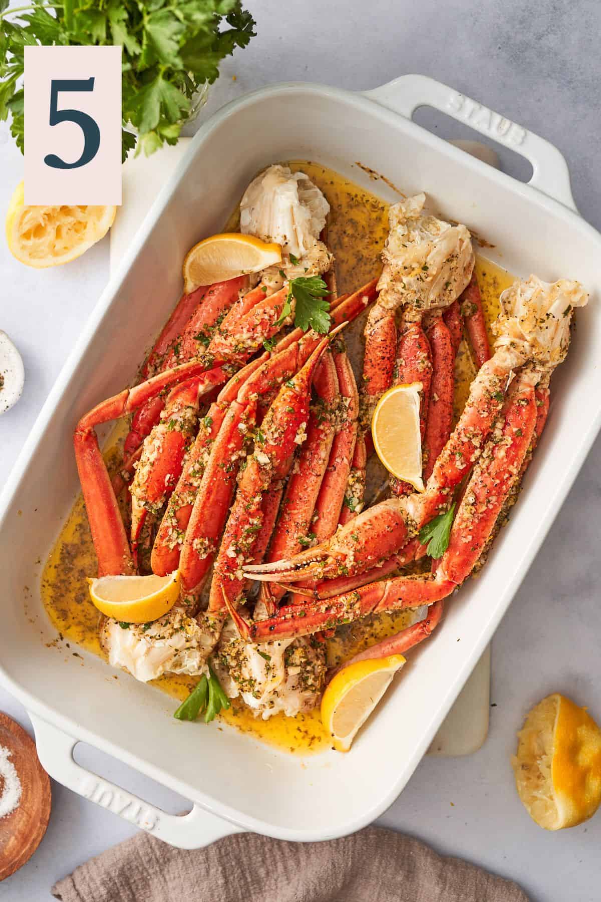 staub casserole dish with baked crab legs smothered in garlic herb dipping sauce, with lemon wedges.