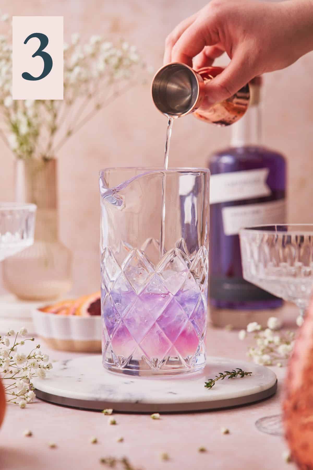 hand pouring in a clear liqueur into a mixing glass with purple gin that begins to change to a light pink color. 