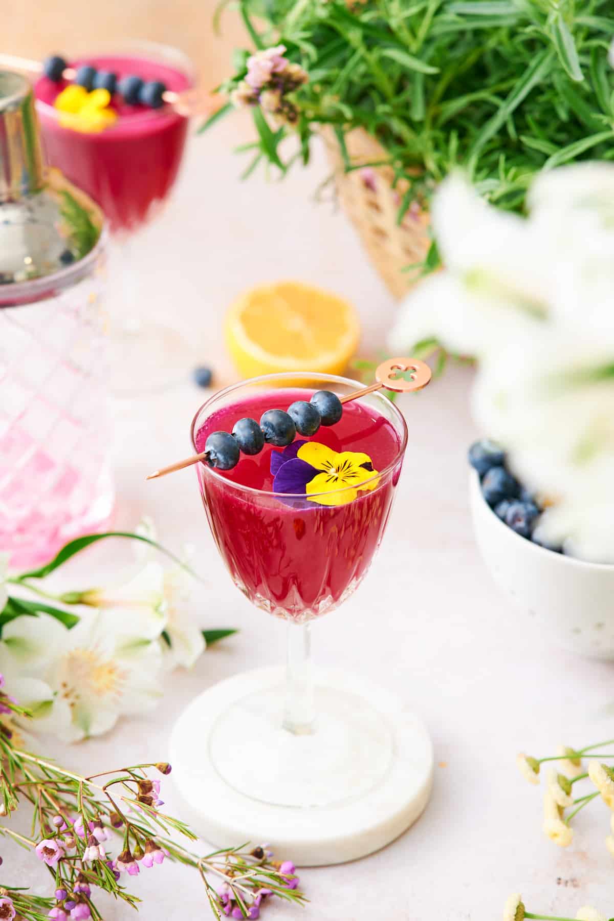 stunning and whimsical blueberry gin cocktail with edible flowers and fresh blueberries, surrounded by flowers, a cocktail shaker, and fresh blueberries. 