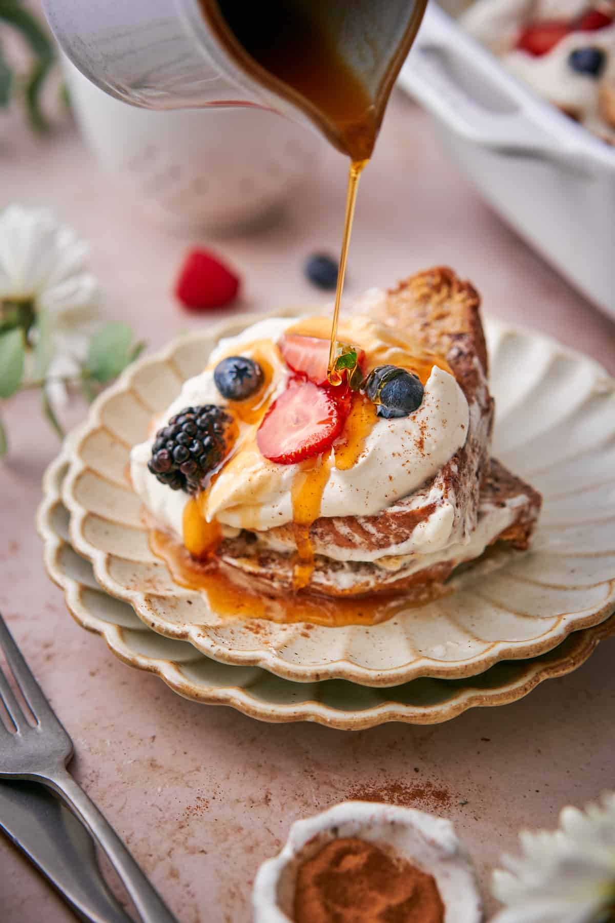 ruffled plates stacked with brioche french toast casserole, garnished with whipped cream, berries, and maple syrup being drizzled on.