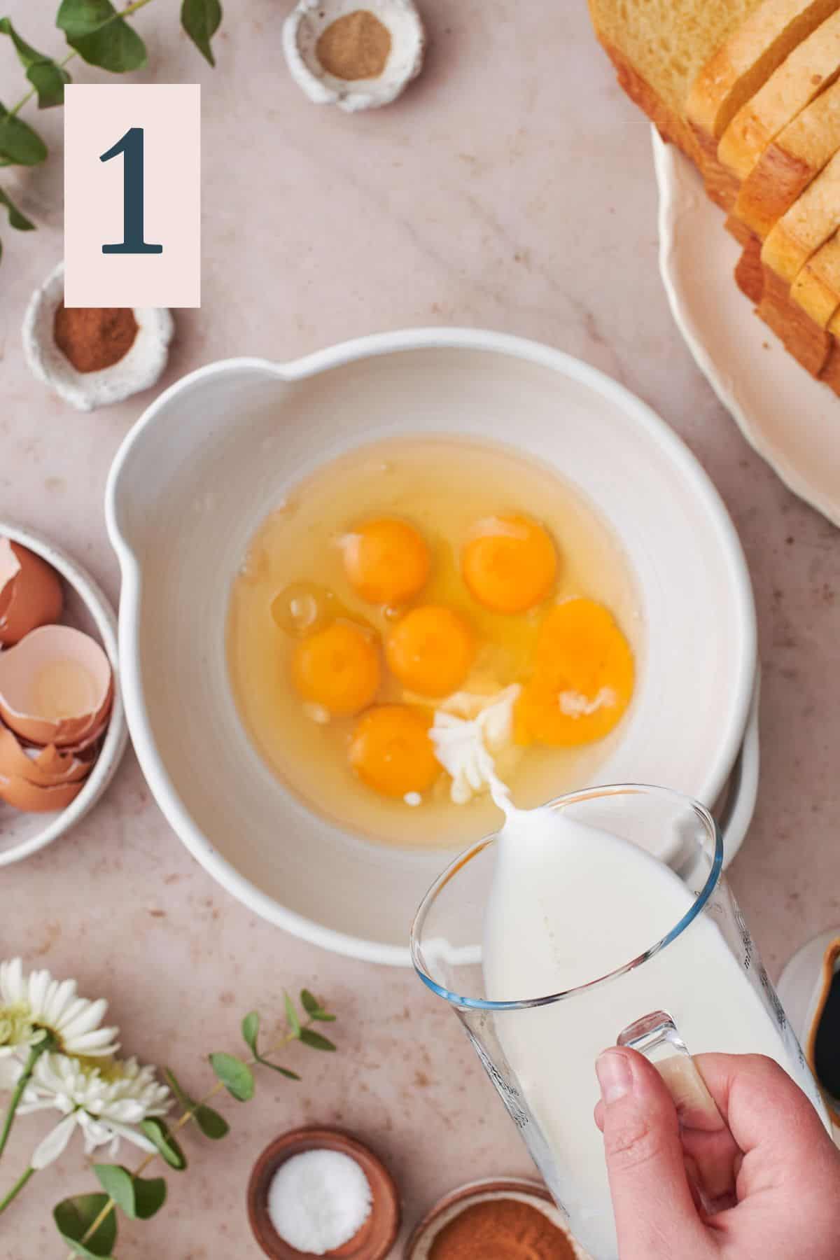 hand pouring milk into a large bowl with whole eggs, surrounded by other ingredients to make the french toast. 