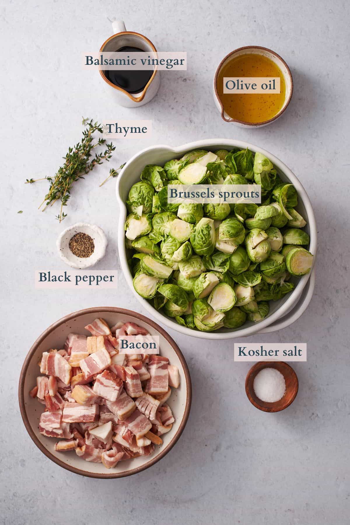 ingredients to make brussels sprouts with bacon and balsamic laid out on a table in ceramic bowls, with text overlaying to denote each ingredient.