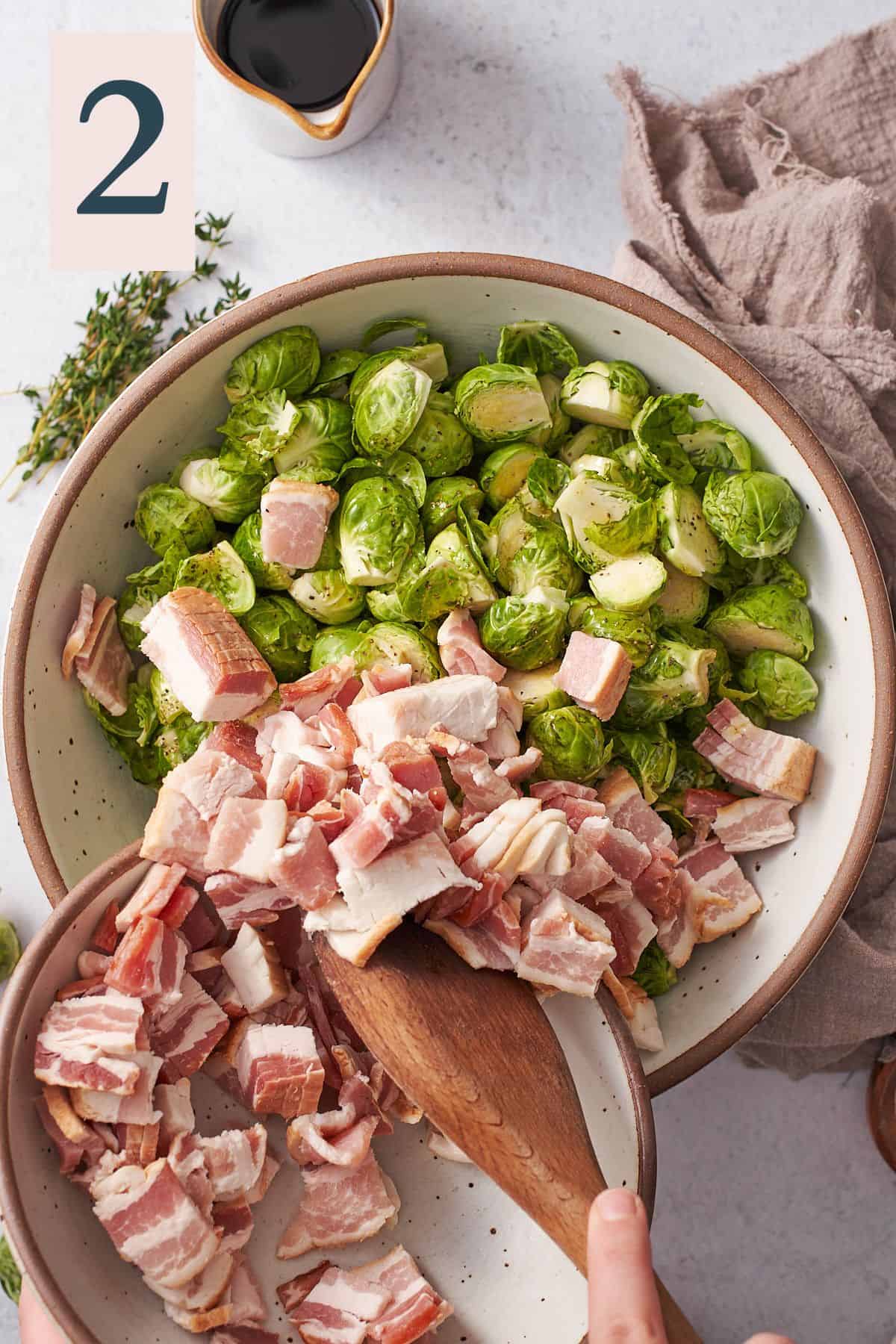hand pushing bacon pieces from a bowl into a bowl of brussel sprouts with a wooden spoon.