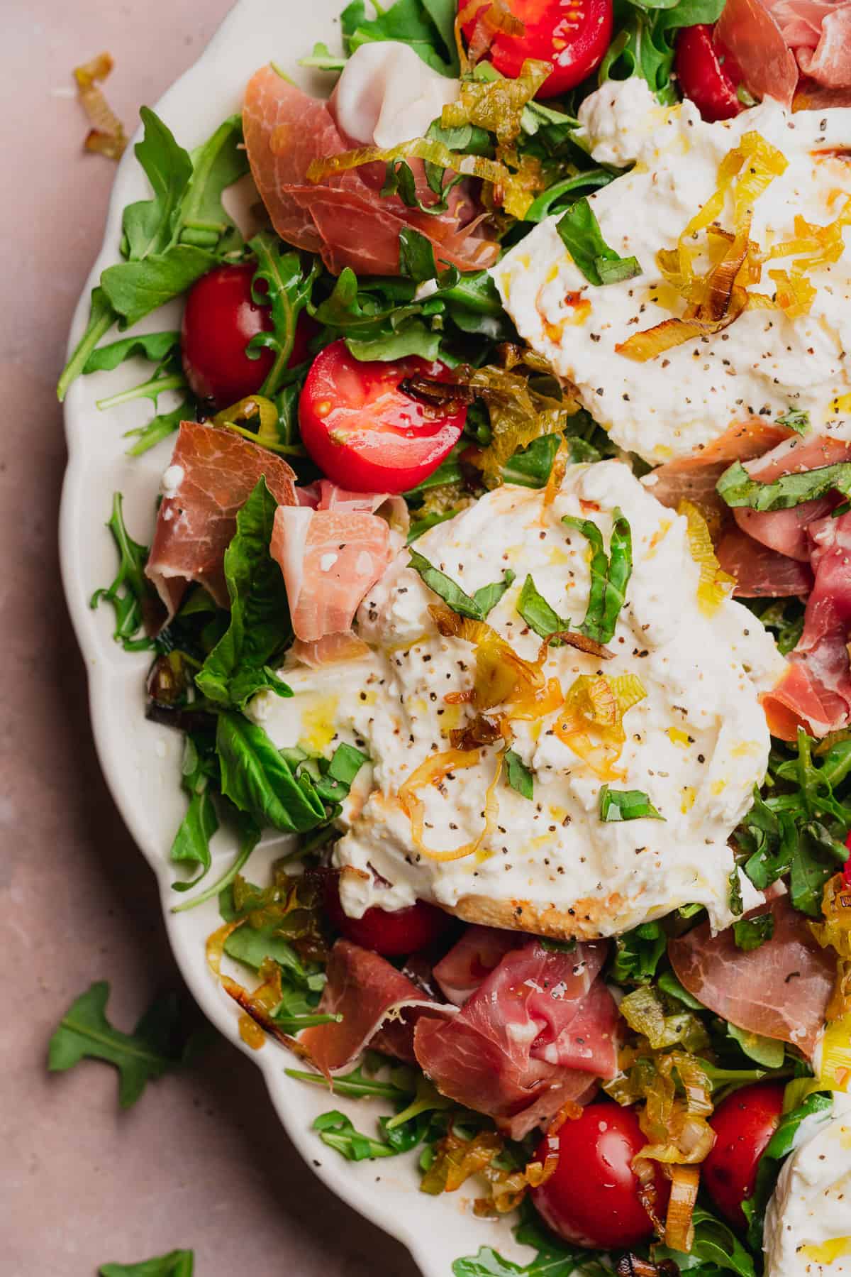 very close up shot of a burrata and prosciutto arugula salad with fresh basil, tomatoes, and fried leeks. 