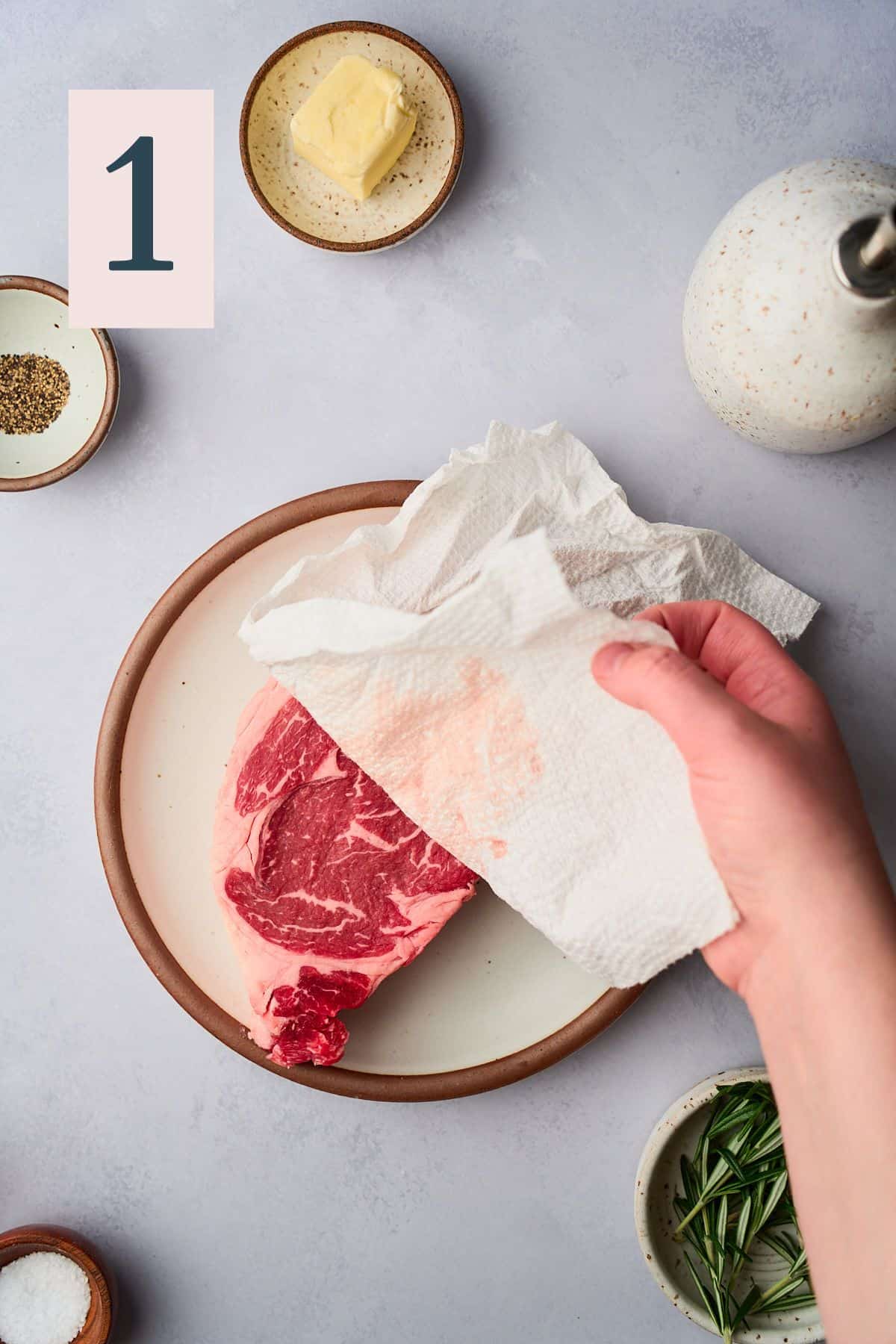 Hand using a paper towel to dry off a large ribeye steak. 