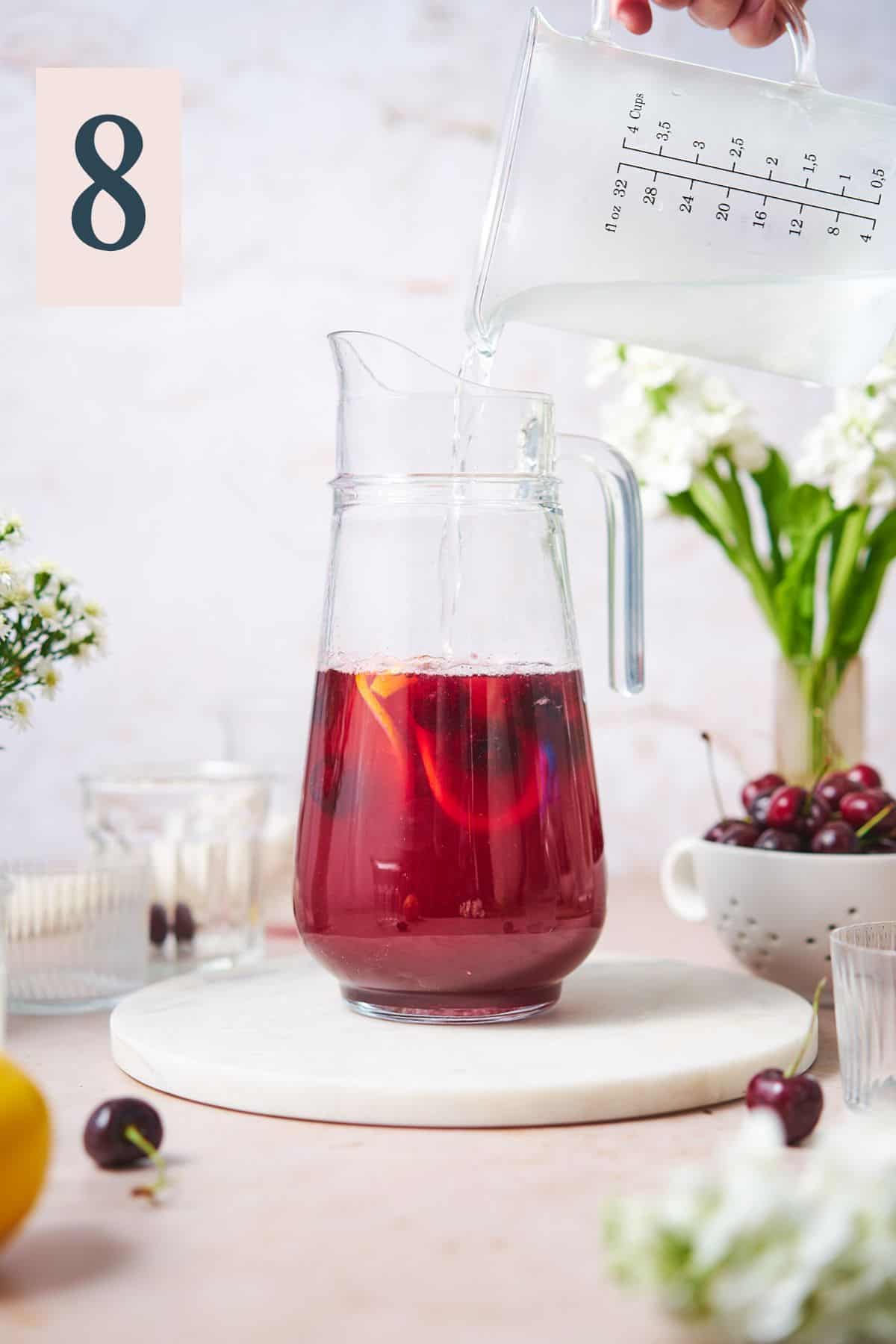 hand pouring in cold water into a large pitcher with cherry syrup and lemon juice. 