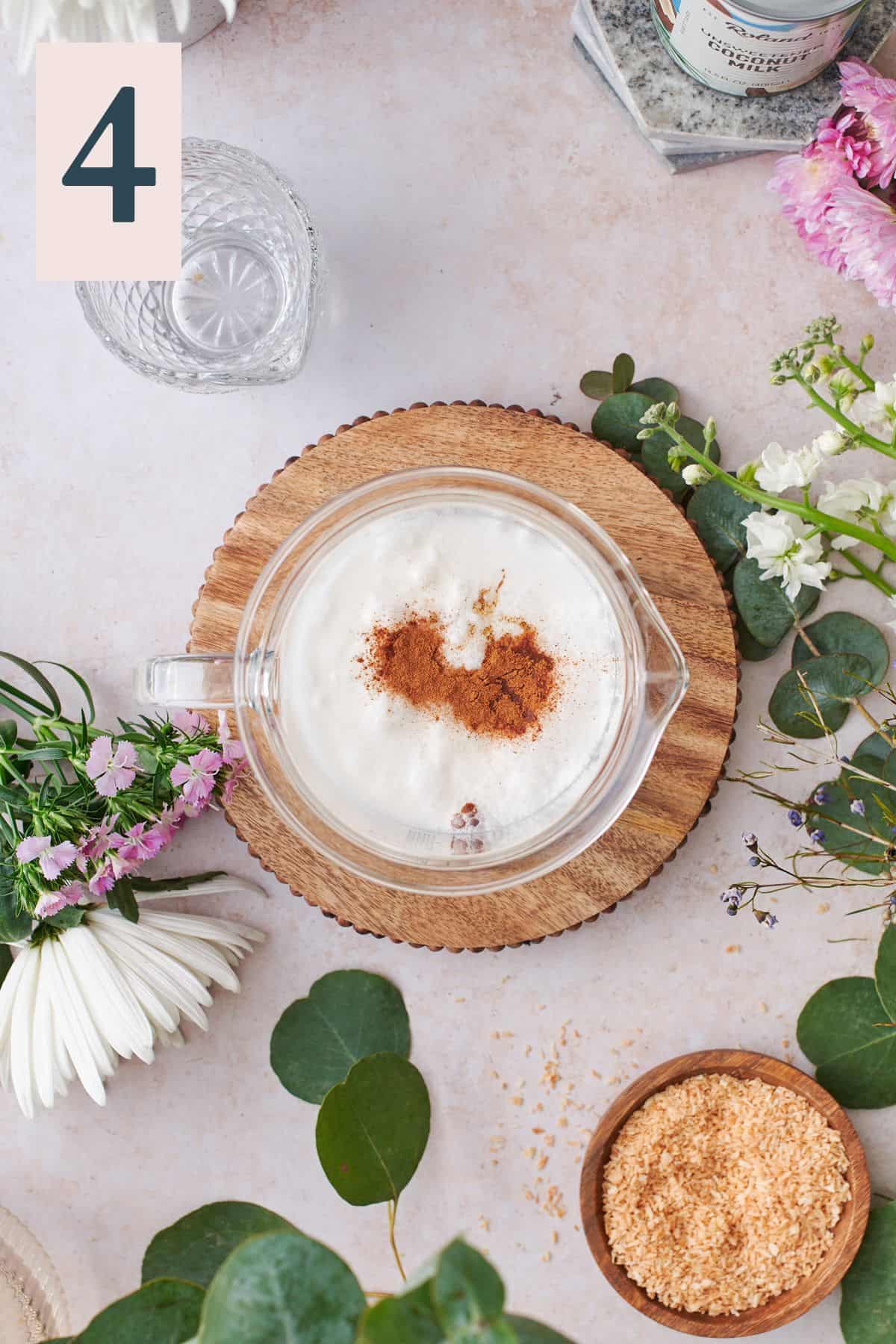 overhead view of coconut milk in a large beaker surrounded by flowers on a wooden platter, with cinnamon, salt, and vanilla extract inside the beaker with the coconut milk. 