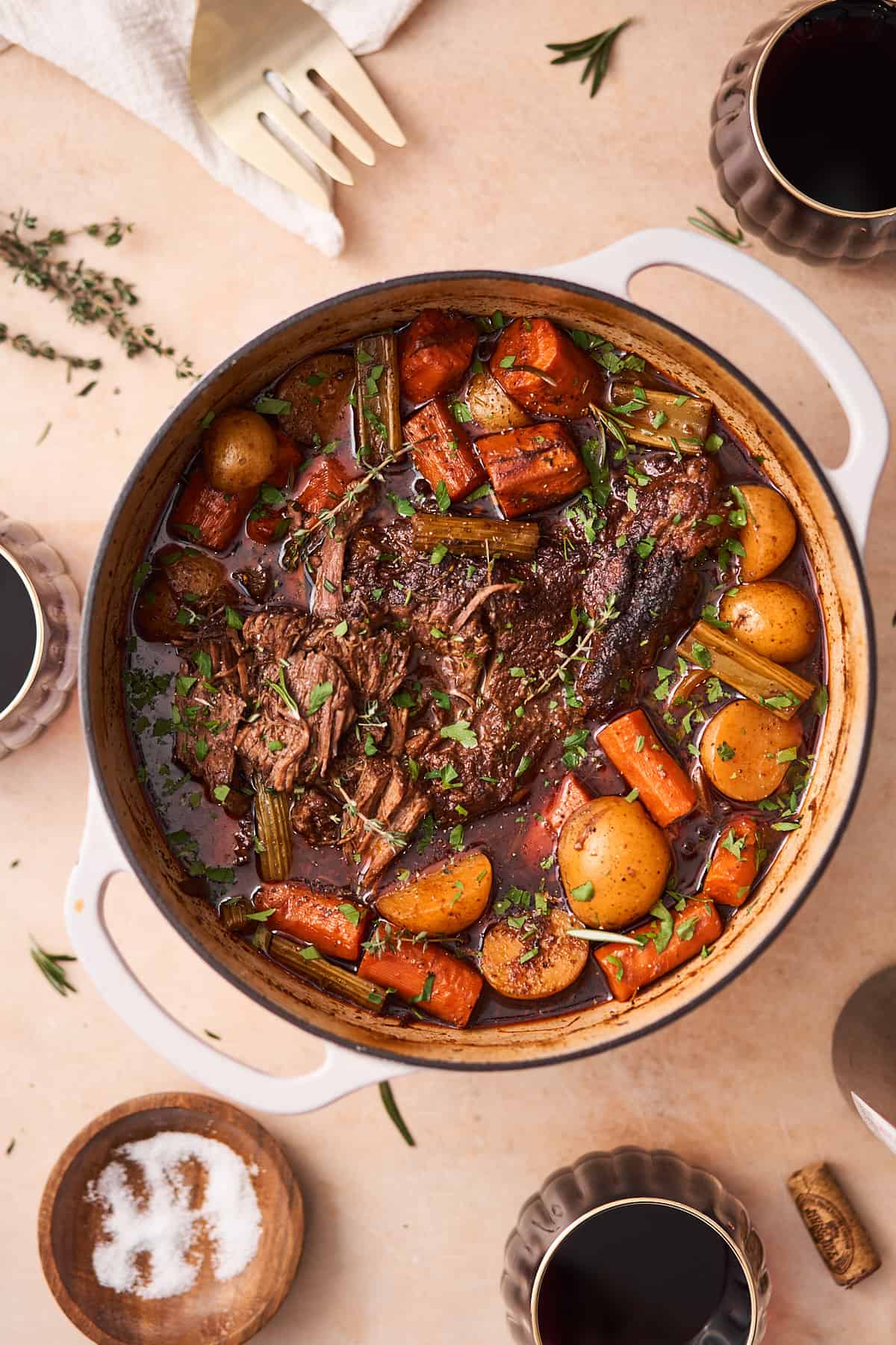 chuck roast in a dutch oven with potatoes, carrots, and celery cooked in a broth, surrounded by thyme, and red wine in glasses.