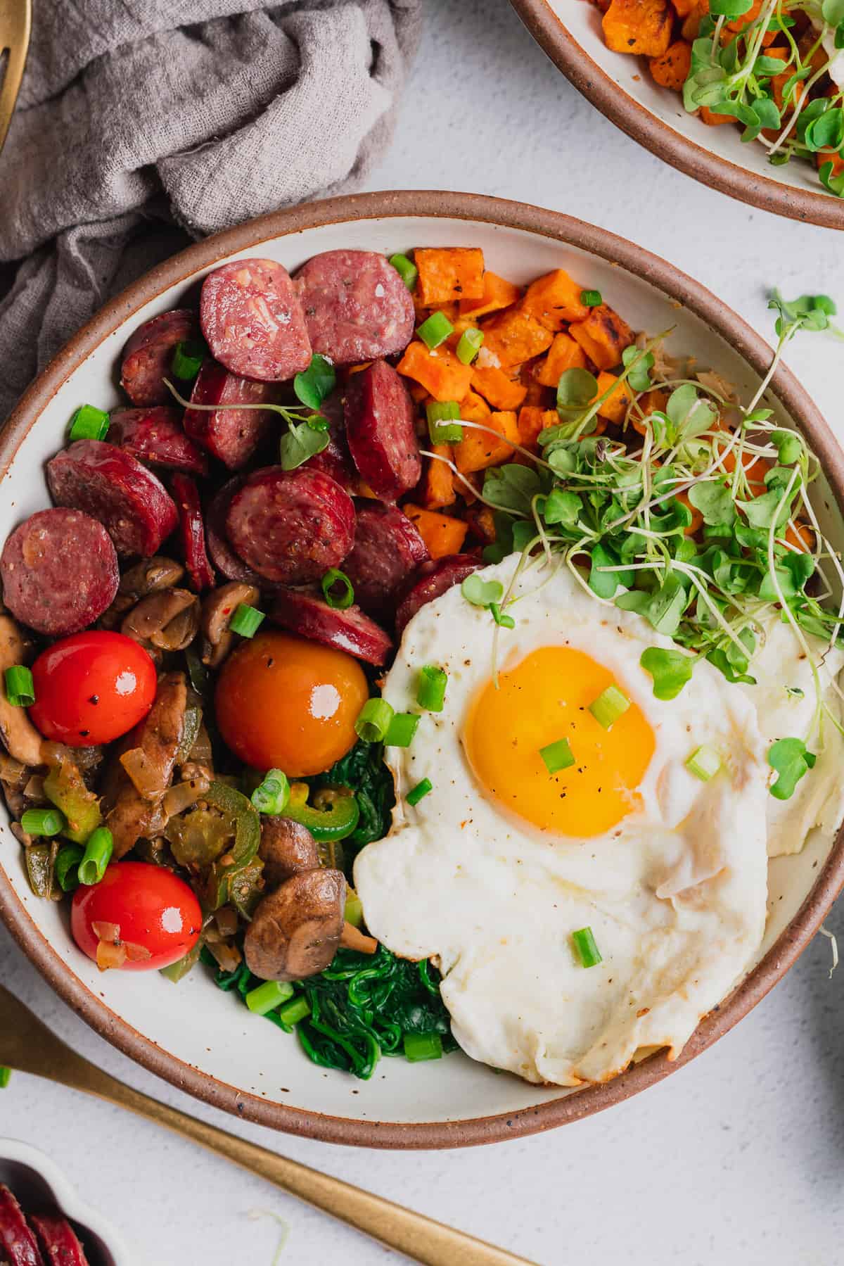 close up shot of an energy bowl with tomatoes, mushrooms, sweet potatoes, green onions, spinach, an egg, smoked sausage, and broccoli greens