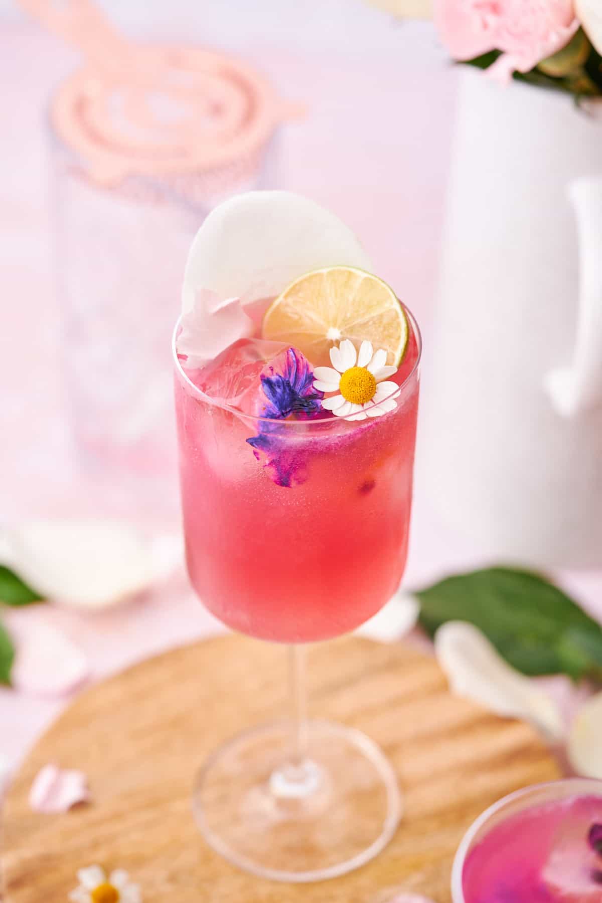 45-degree shot down of a bright pink floradora cocktail, with a wooden round trivet underneath, a mixing glass behind it, a white ceramic vase, and flower petals. 
