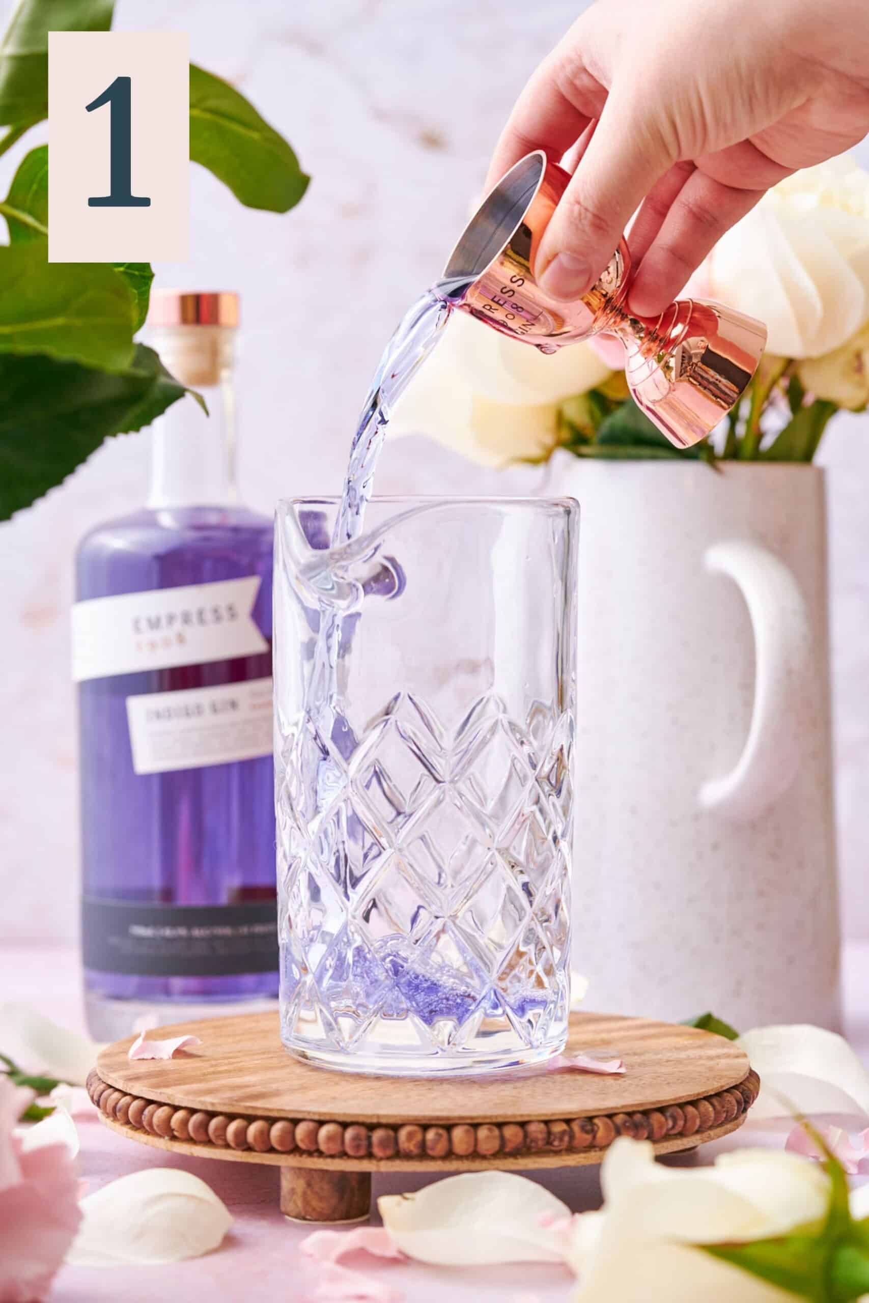 hand pouring purple empress gin into a cocktail mixing glass, with white roses and pink carnations in the background. 