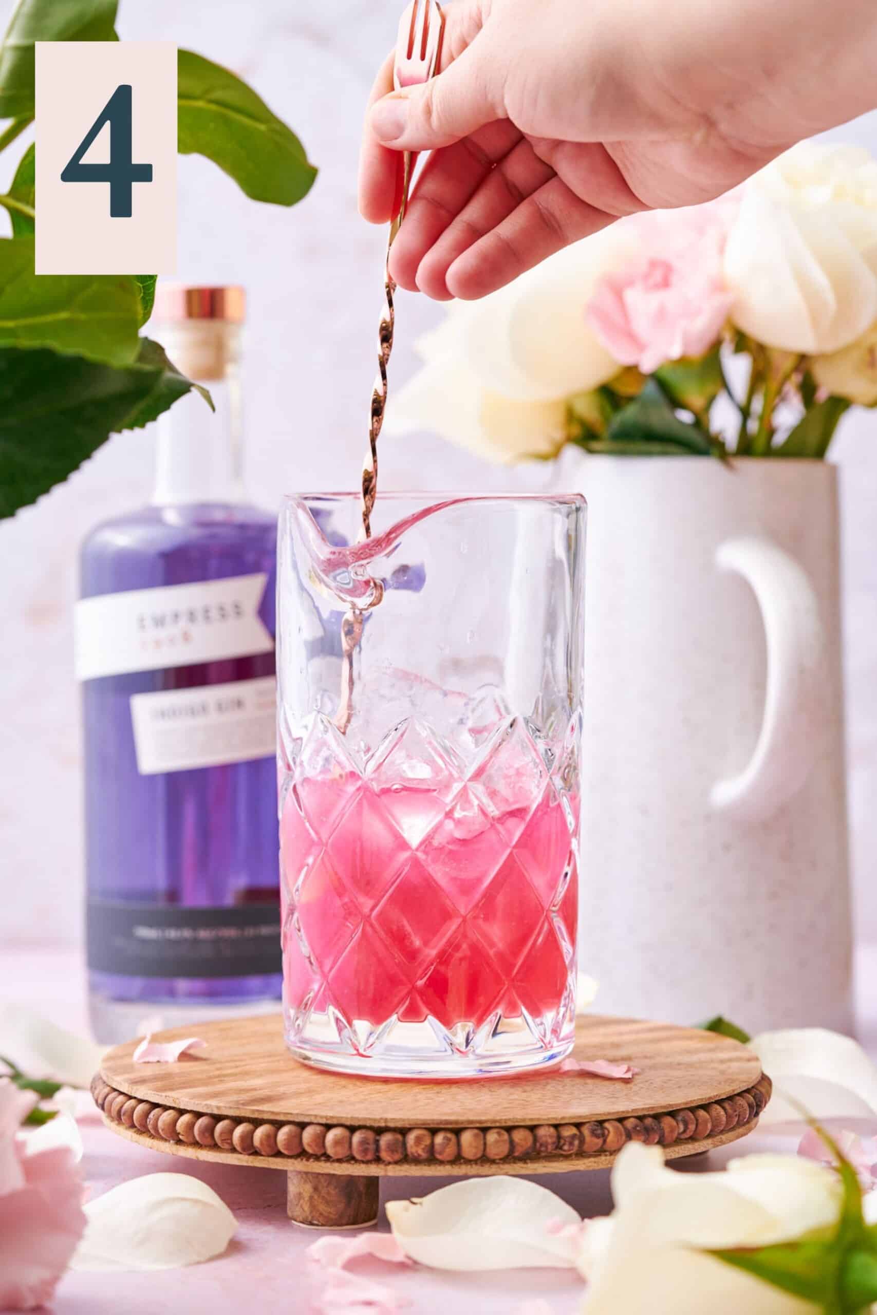 hand stirring a bright pink cocktail in a mixing glass with a bar spoon, and flowers in the background. 