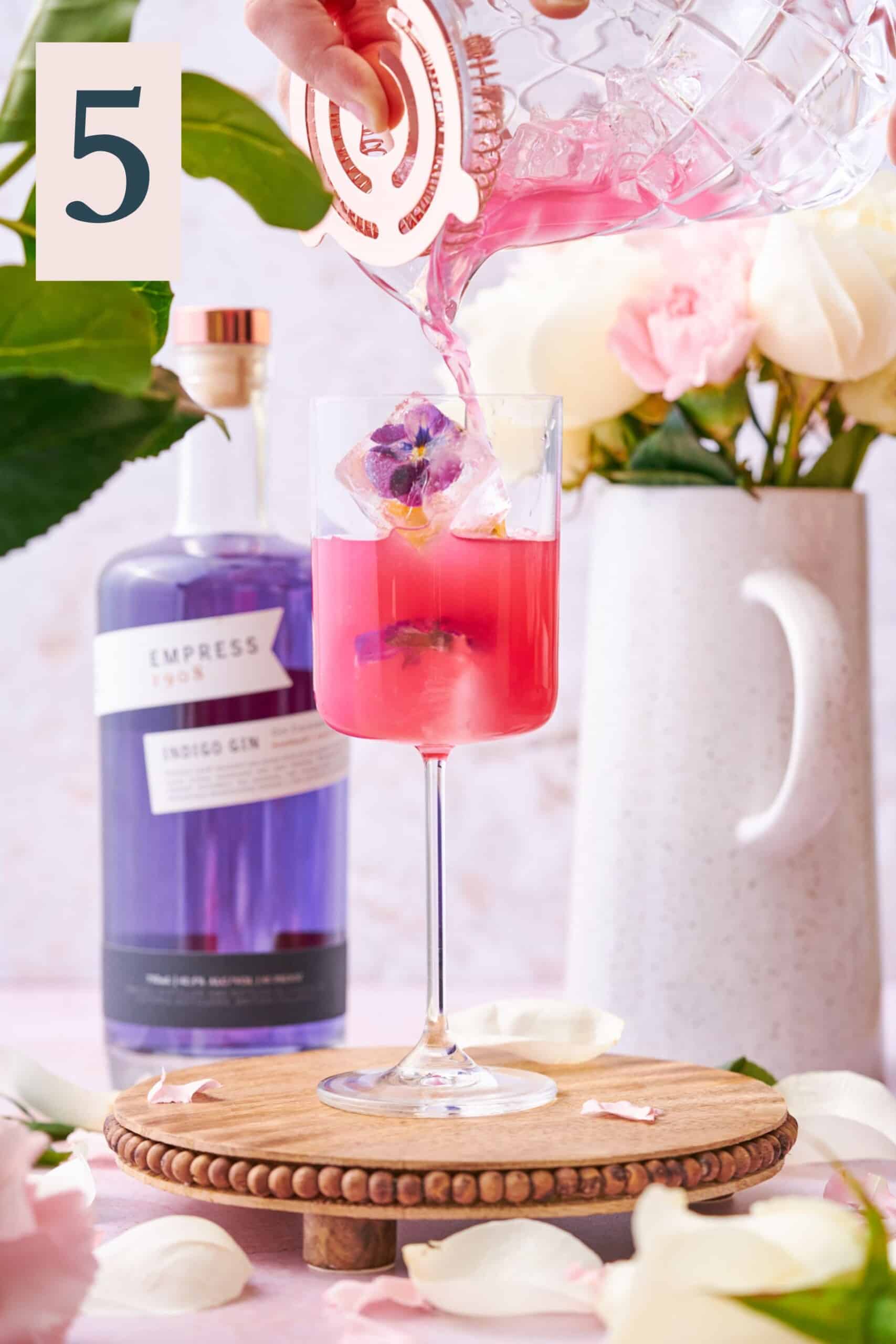 hand pouring bright pink liquid into a glass with floral ice cubes, and Empress gin in the background. 
