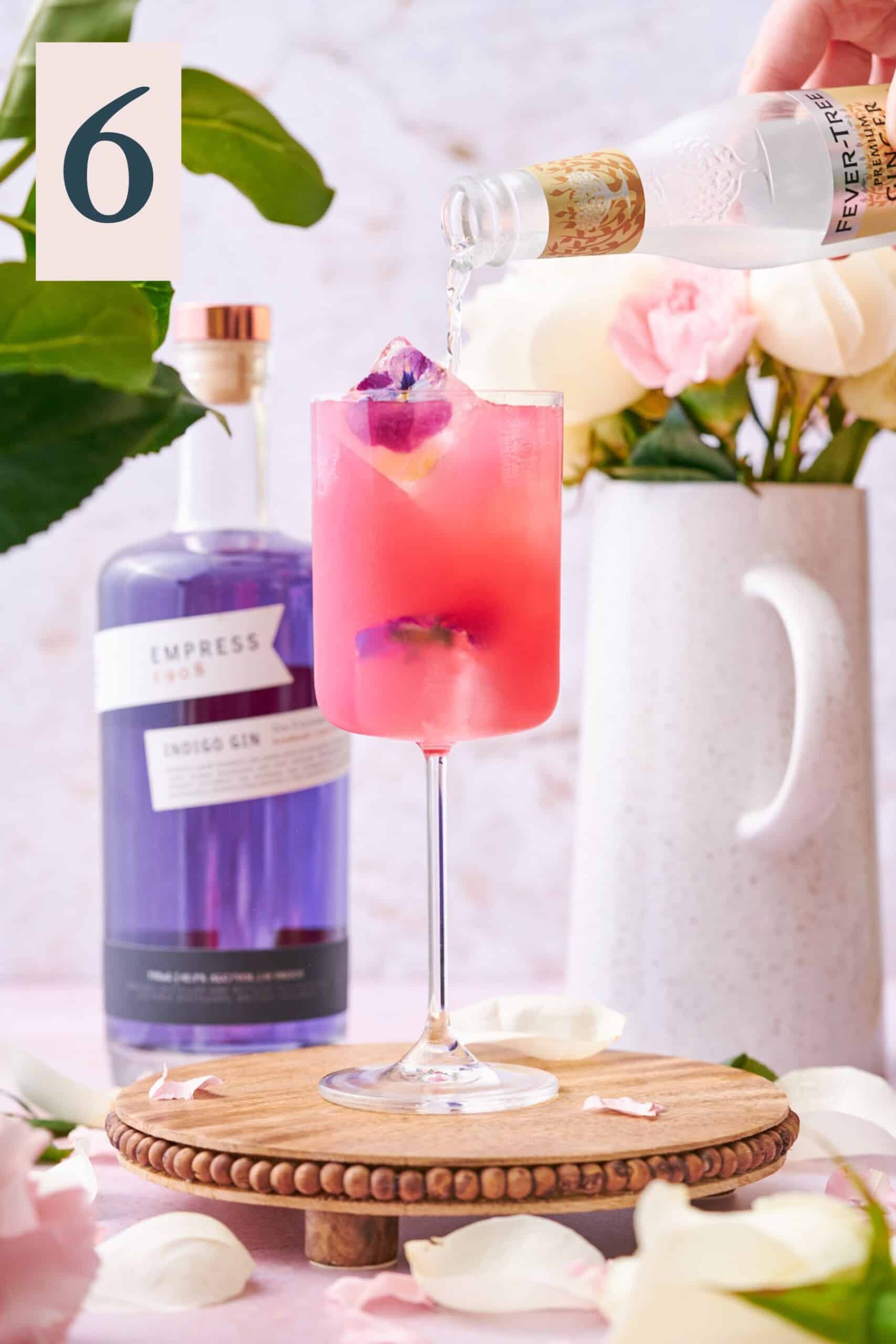 pouring ginger beer into a bright pink Floradora cocktail, with a bottle of indigo Empress gin in the background, and white and pink flowers.