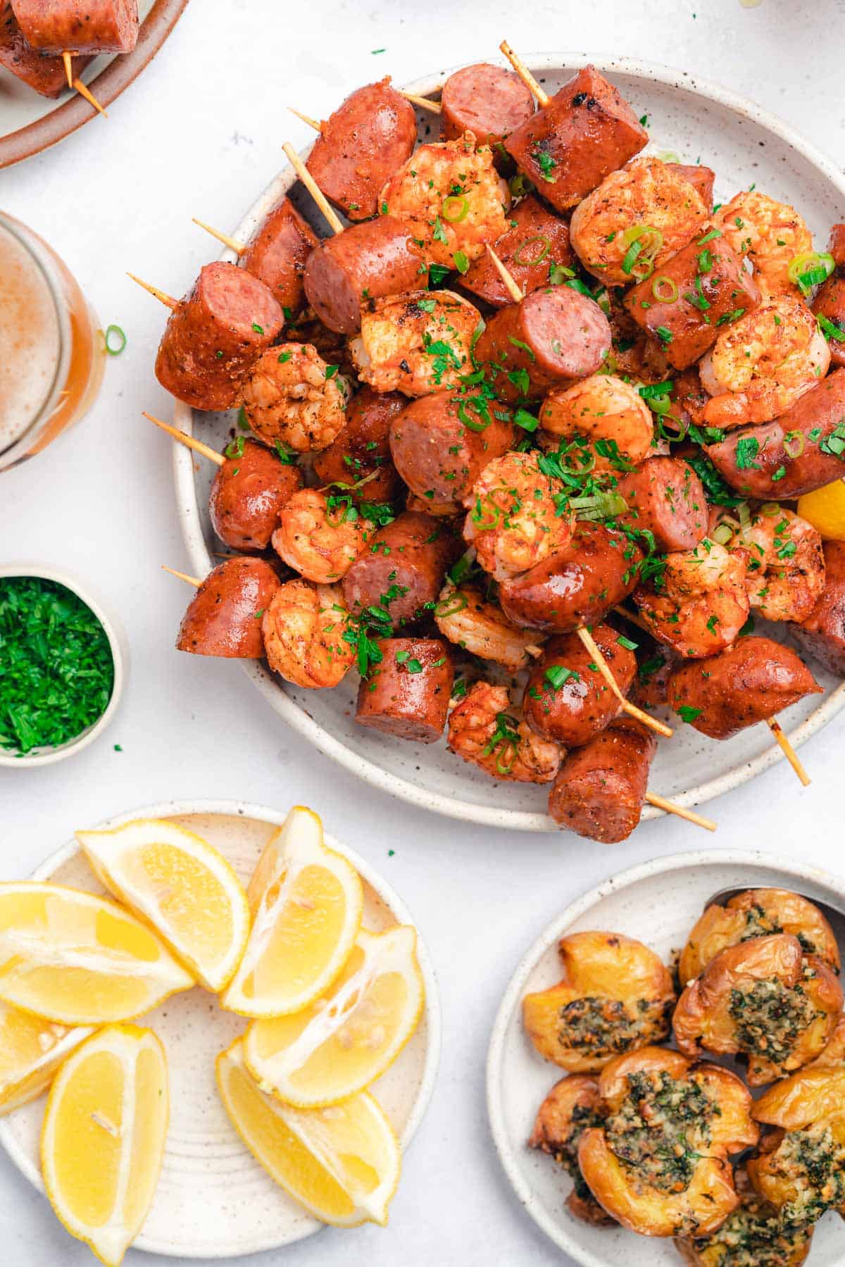 flat lay of grilled smoked sausage and shrimp skewers served with beer, crispy herbed potatoes, lemon wedges and herbs