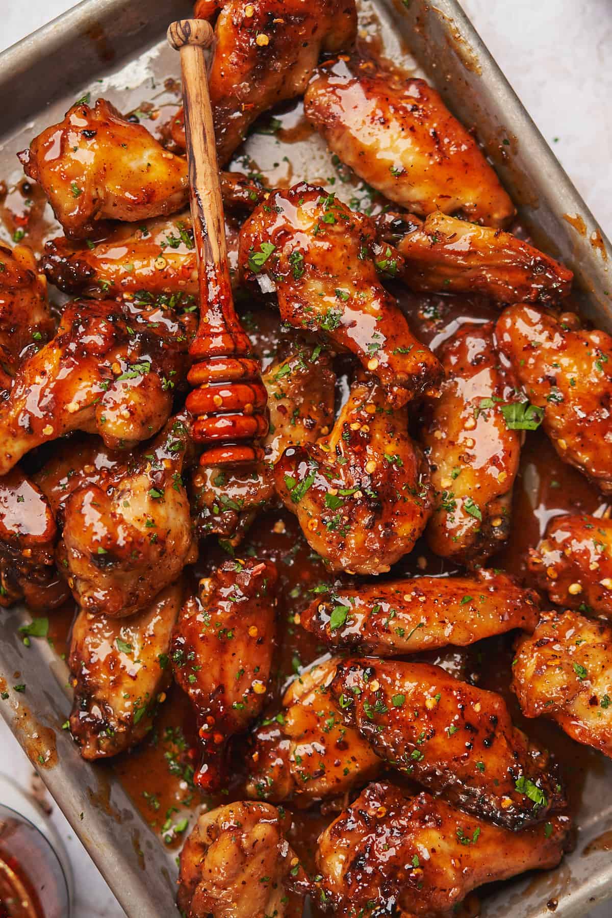 close up shot of honey hot wings in a baking sheet, with a wooden honey dripper putting more spicy honey on top.