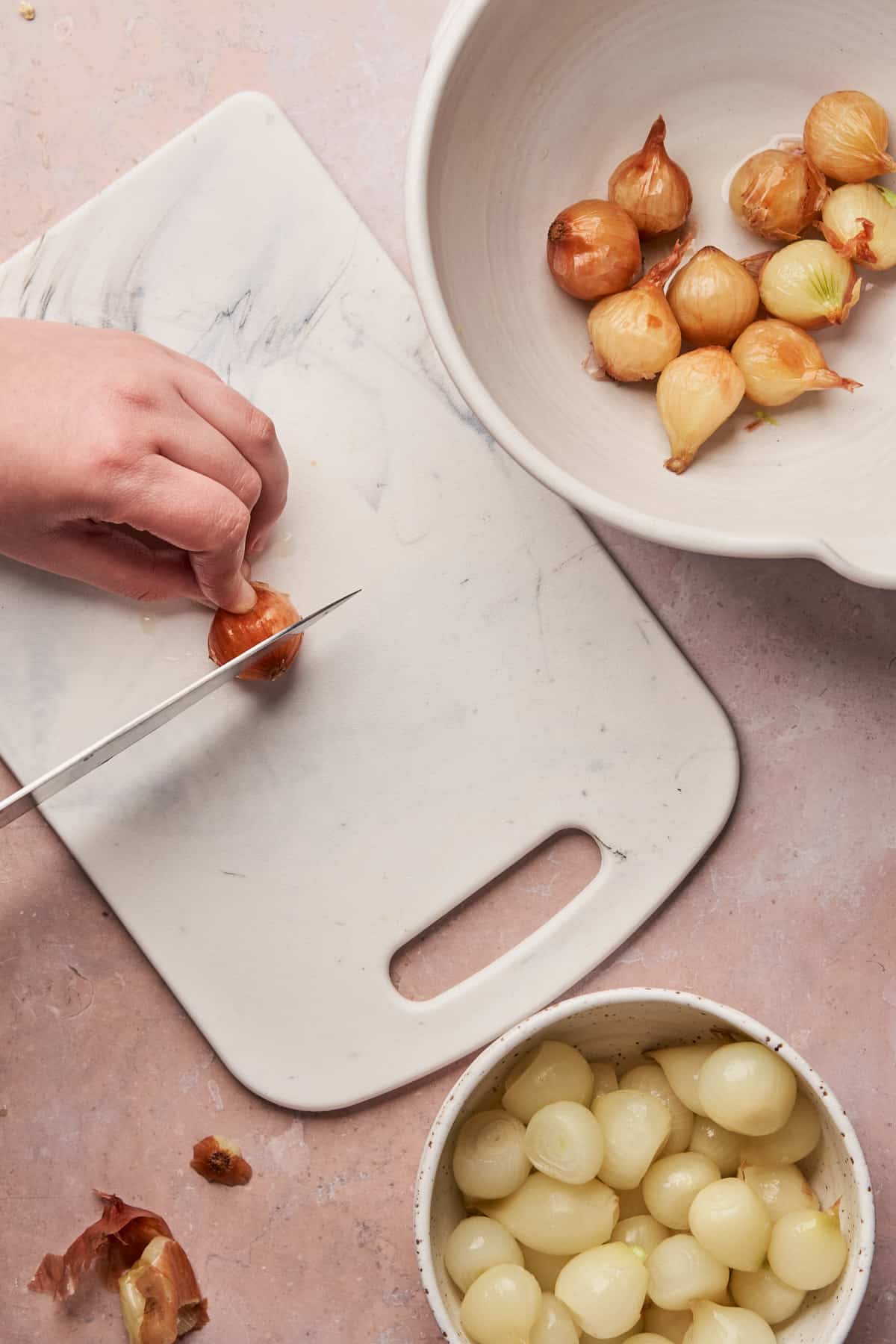 hand cutting off the end of a pearl onion on a cutting board with a knife. 