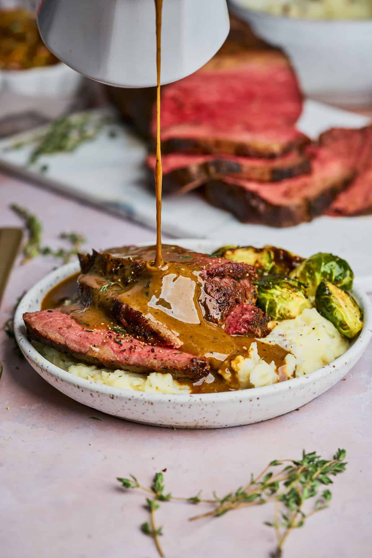 drizzling brown gravy overtop of roast beef on a plate with mashed potatoes and brussel sprouts, with the carved roast beef in the back.