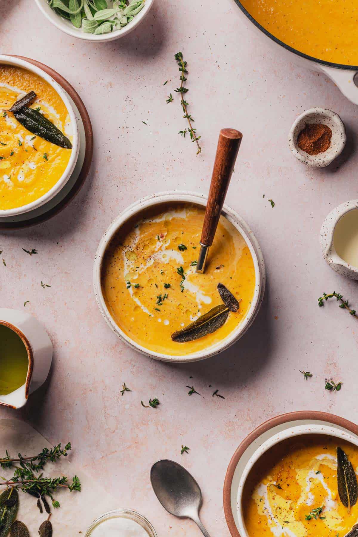 beautiful flay lay shot of keto butternut squash soup surrounded by other full bowls, heavy cream, fresh thyme, nutmeg, salt, pepper and nutmeg