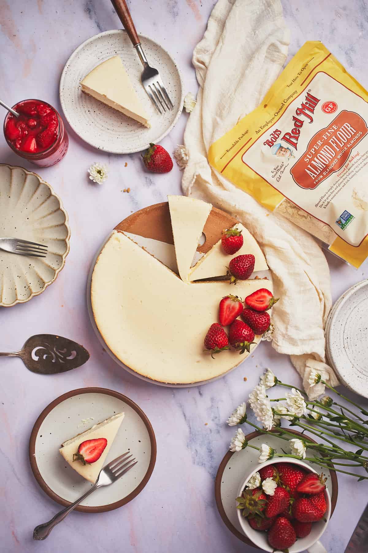 flat lay of a keto cheesecake recipe topped with fresh strawberries, flowers, a bag of Bob's Red Mill Almond flour, and plates with forks and fresh slices of cheesecake on them. 