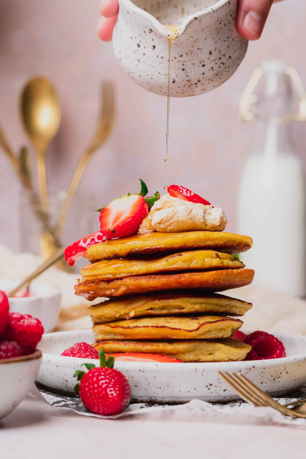 straight on shot of a stack of coconut flour pancakes topped with whipped butter and berries, with maple syrup being poured on 