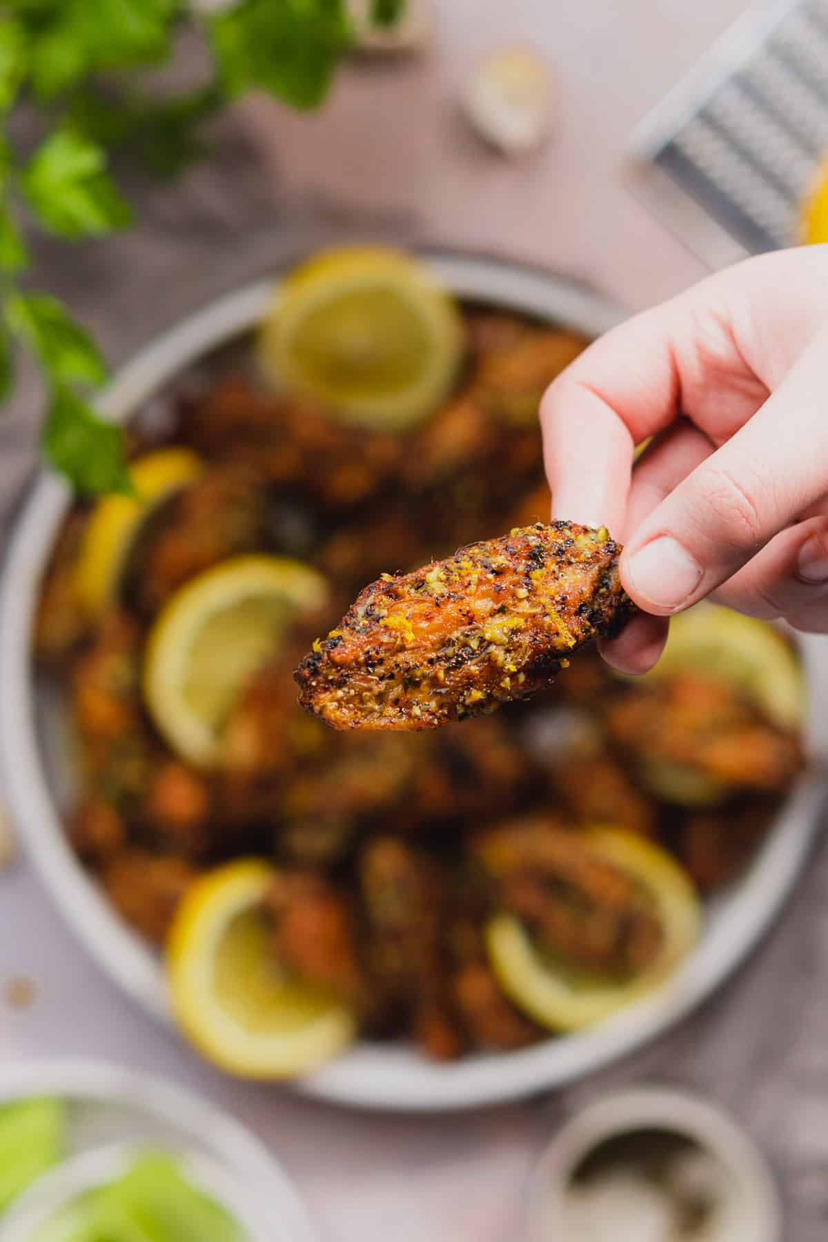 hand holding a lemon pepper wing over a tray of lemon pepper chicken wings with lemon rounds. 