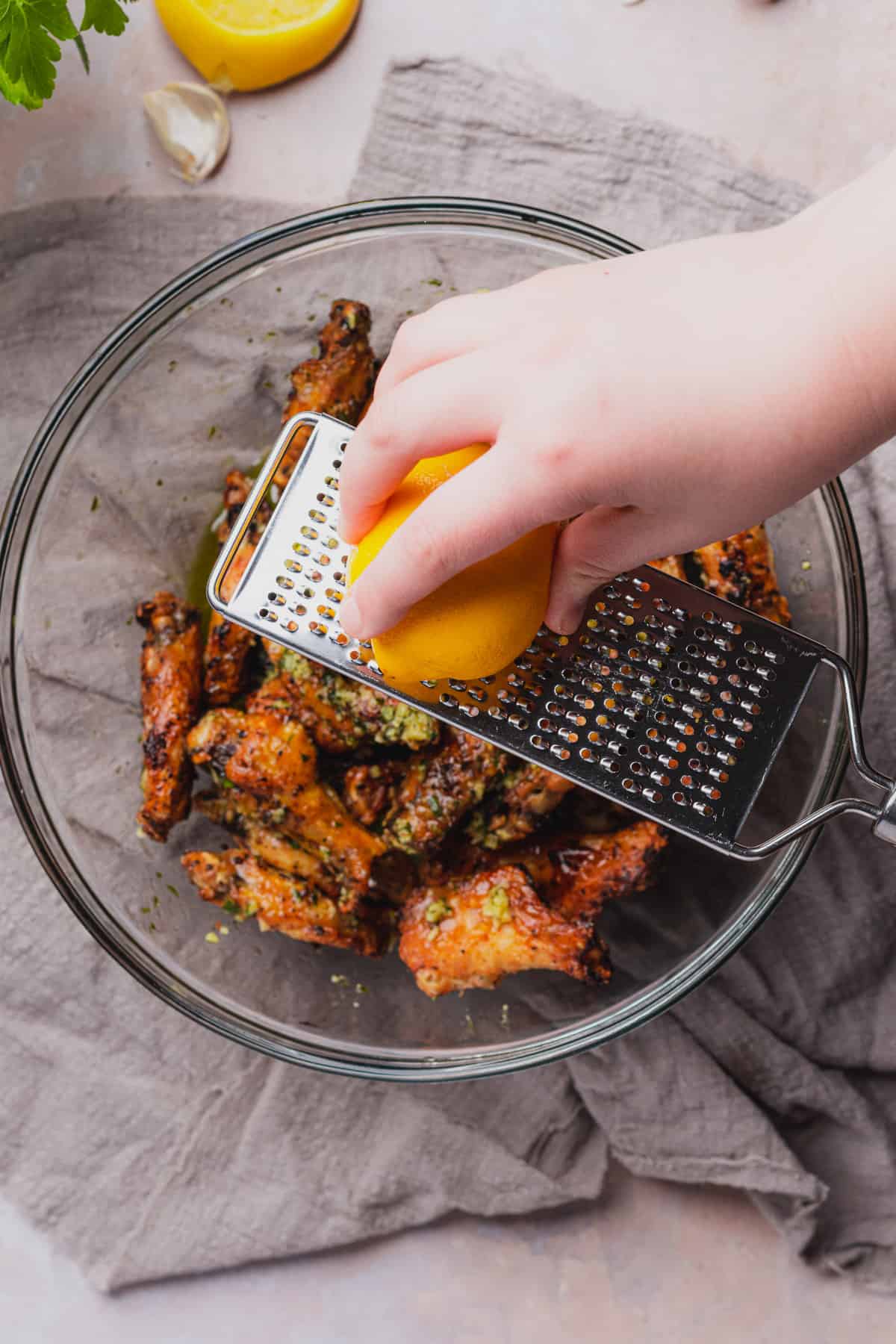 hand zesting a lemon overtop of crispy cooked chicken wings in a large mixing bowl. 
