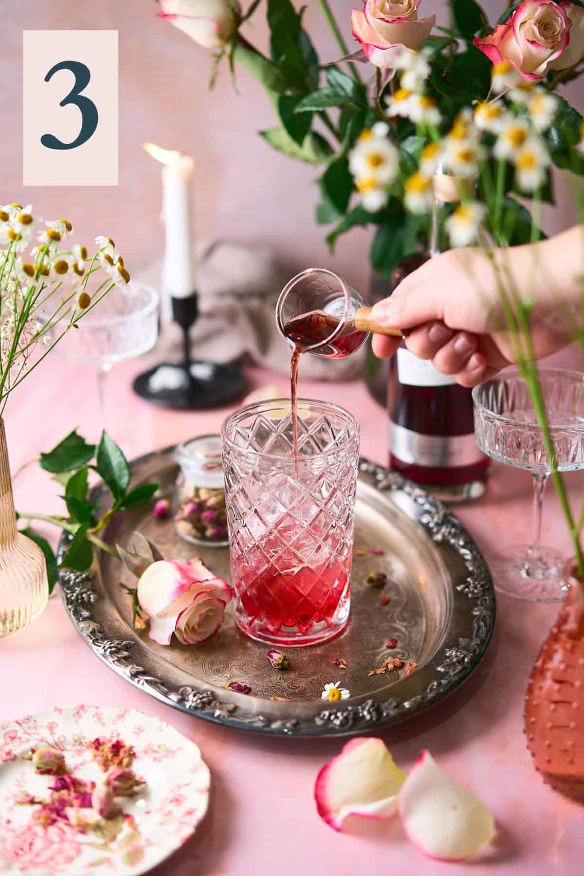 hand pouring raspberry liqueur into a beautiful glass cocktail shaker with an intricate romantic scene of fresh roses, candles and flowers surrounding it.