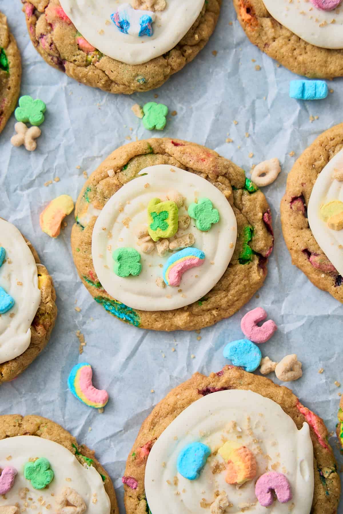 Close of shot of Lucky Charms cookies with marshmallow frosting, and topped with sprinkles, lucky charms marshmallows, and cereal pieces on white parchment paper. 