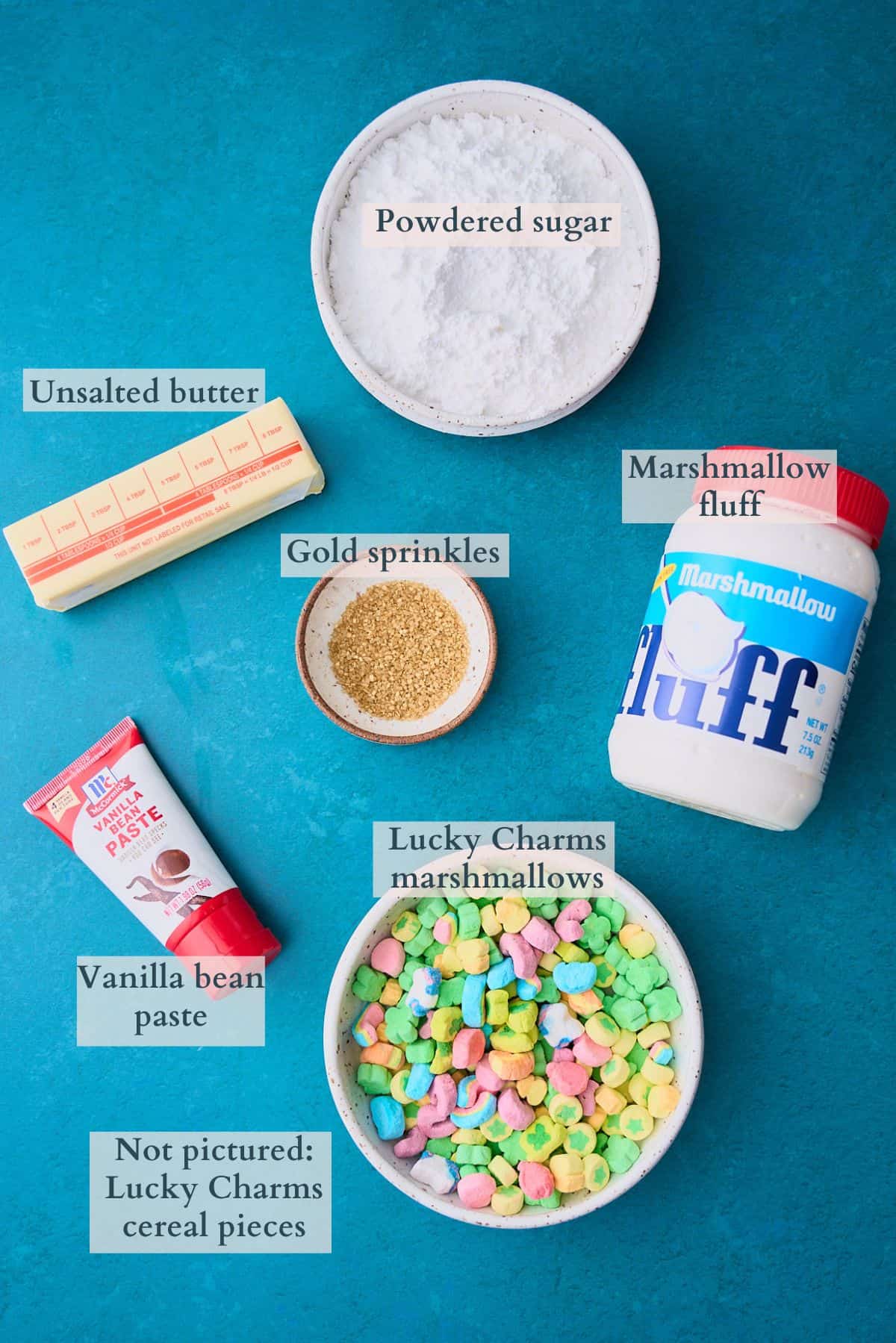 Ingredients laid out on a deep green backdrop to make marshmallow fluff frosting with Lucky Charms marshmallows and labeled to denote each ingredient. 