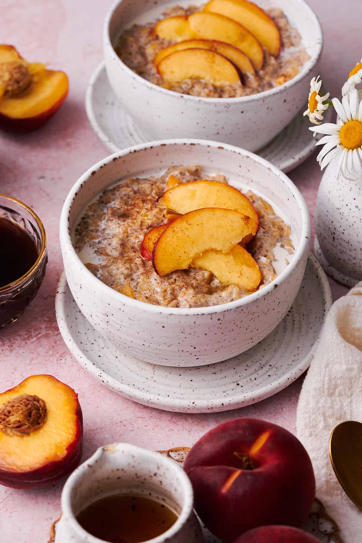 45-degree shot of a bowl of peach oatmeal on a plate surrounded by coffee, flowers, and fresh peaches. 