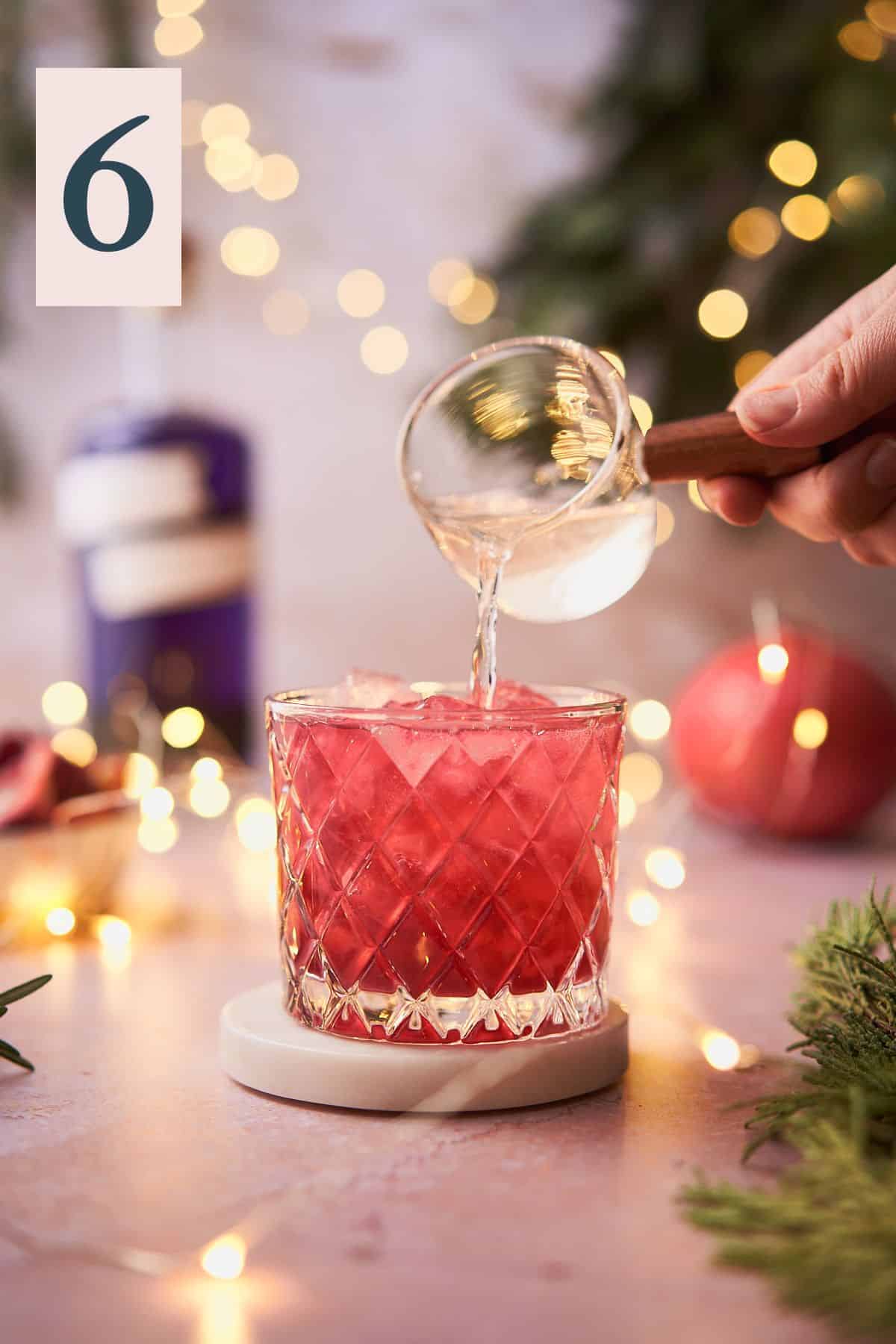 hand pouring ginger beer to top a bright red cocktail in a low ball glass, with twinkling lights, pomegranate, and balsam in the background.