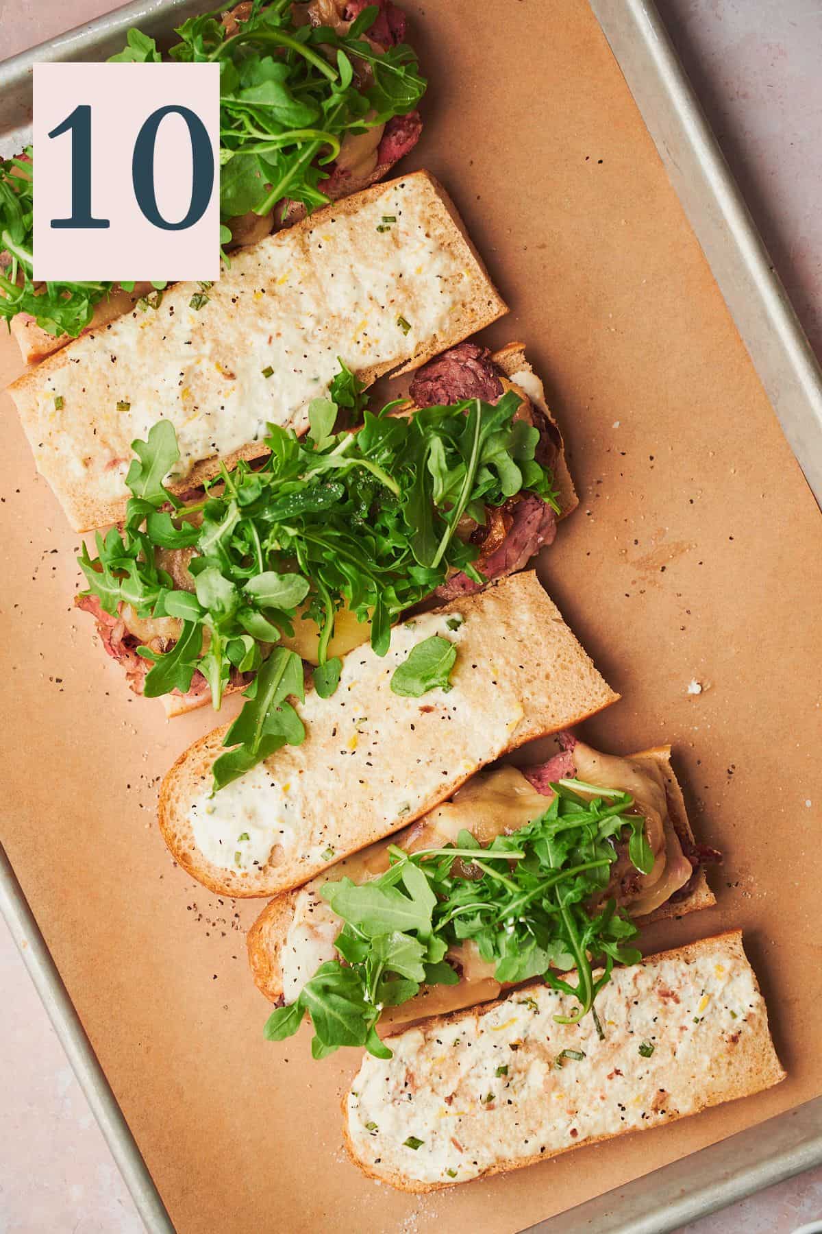 baguettes full of roast beef, cheese, and arugula open and facing the camera on a baking sheet. 