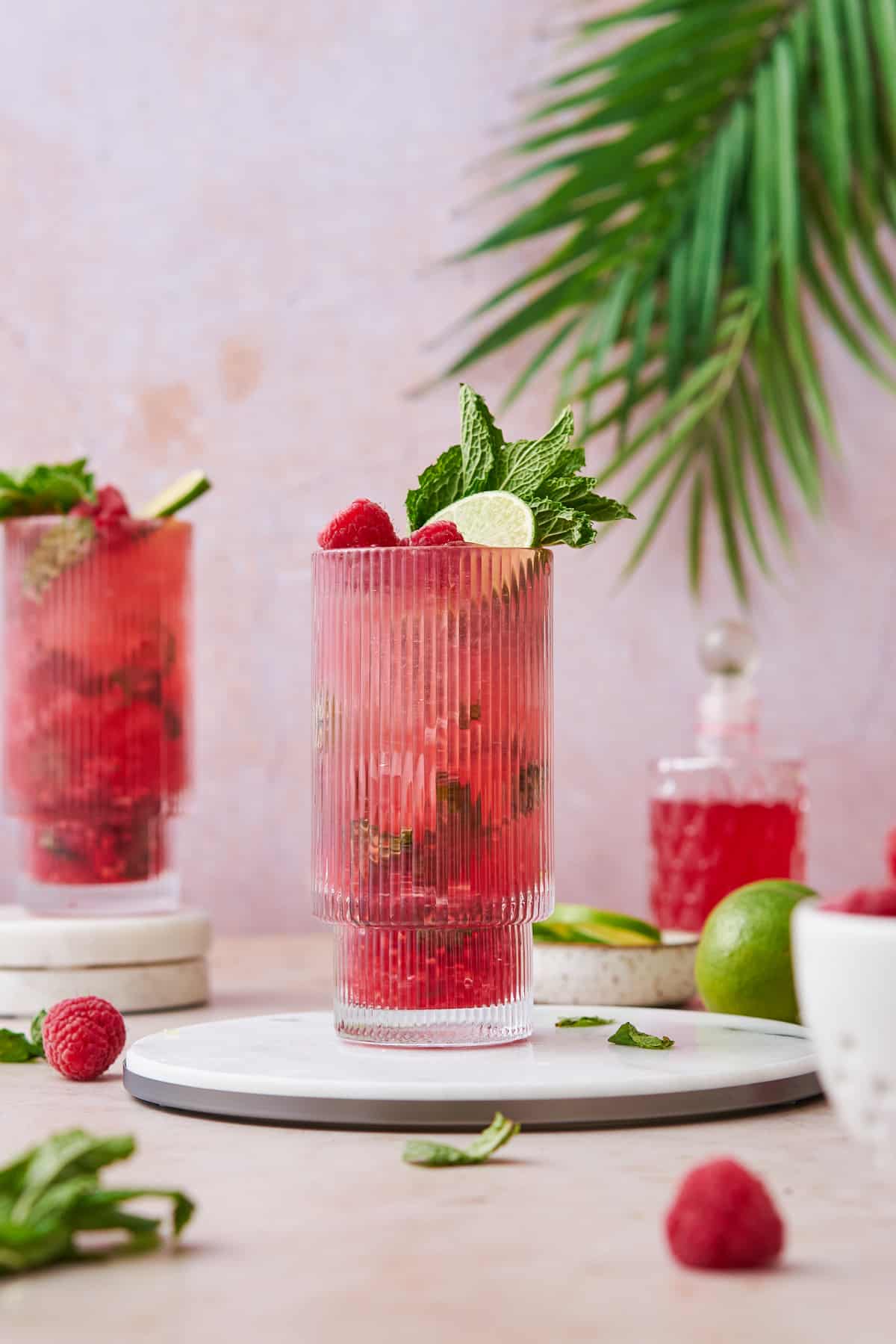 Red raspberry mojito garnished with mint, lime, and raspberries with a pink background and tropical plants in the back.