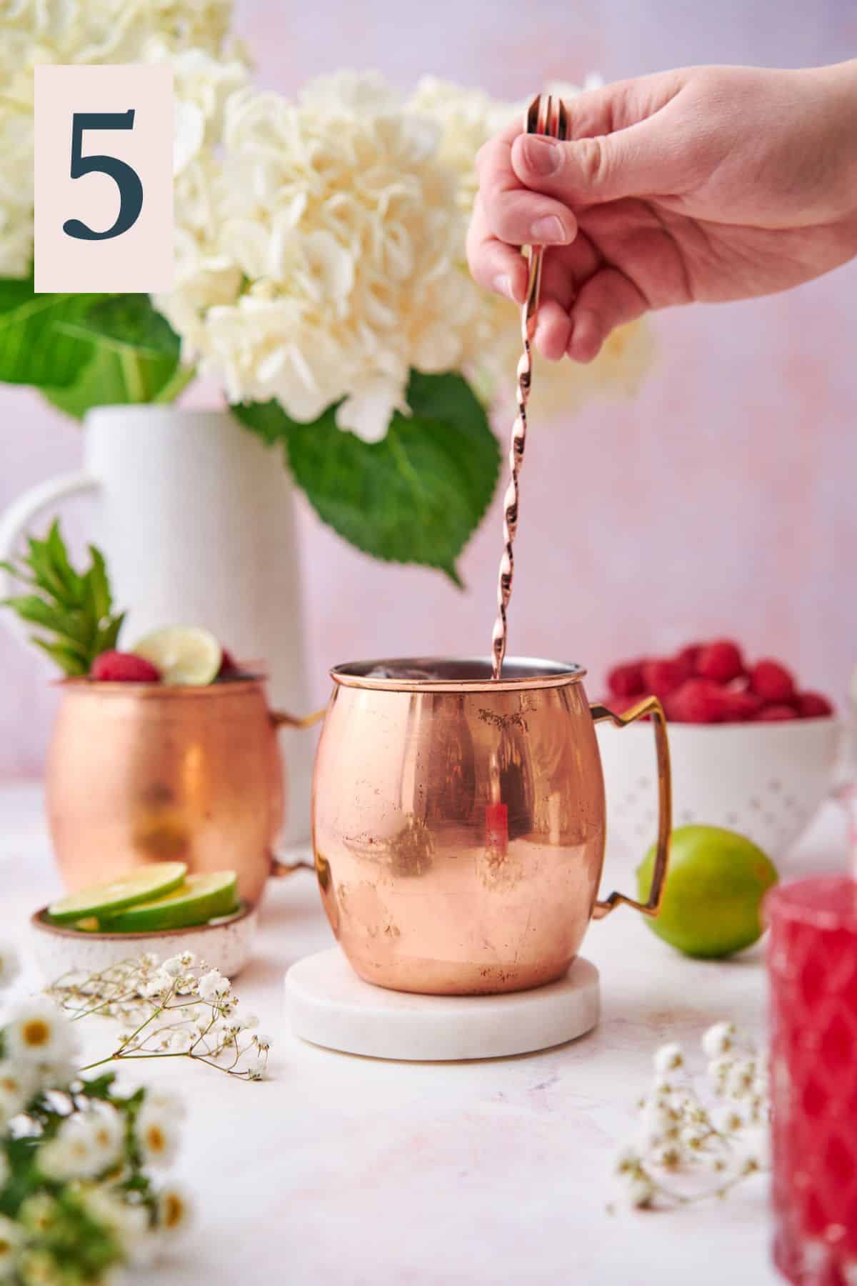 hand stirring a cocktail in a copper moscow mule mug with fresh raspberries, hydrangeas, and other small white flowers.  