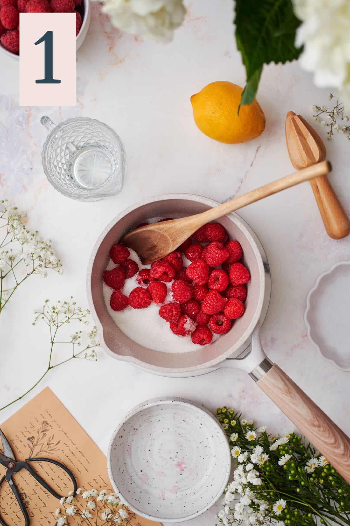 raspberries and sugar in a small sauce pan, surrounded by glassware, flowers, lemon, and water.