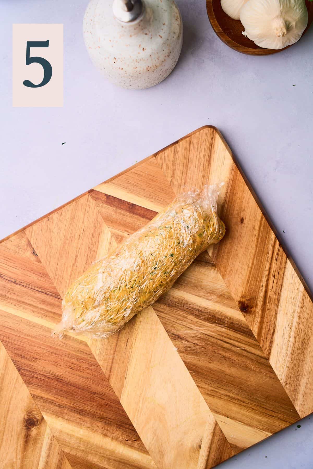 Log of compound butter rolled up in plastic wrap on a wooden cutting board. 
