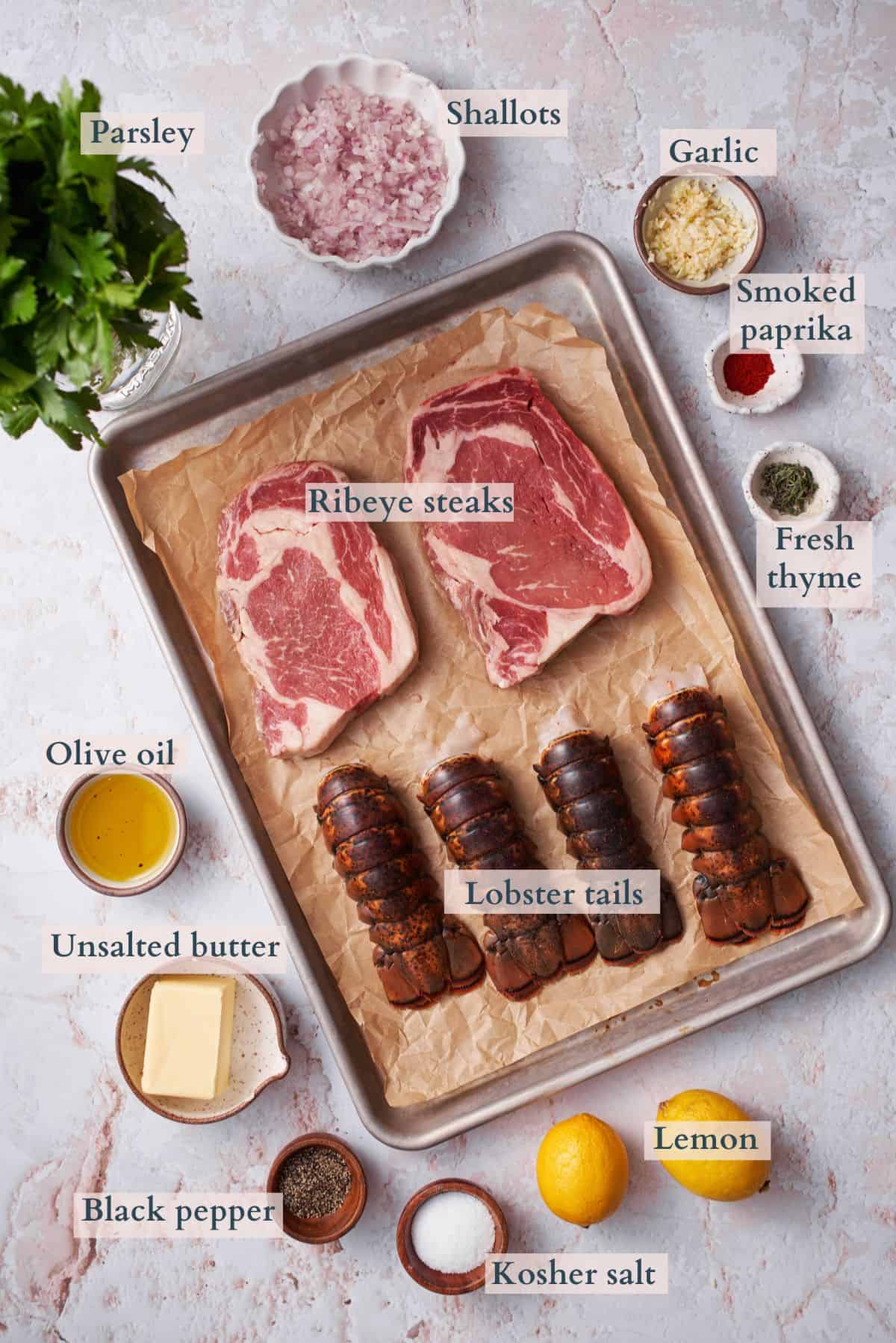 ingredients to make steak and lobster surf and turf laid out on a baking sheet and in bowls, with labels to denote each ingredient. 